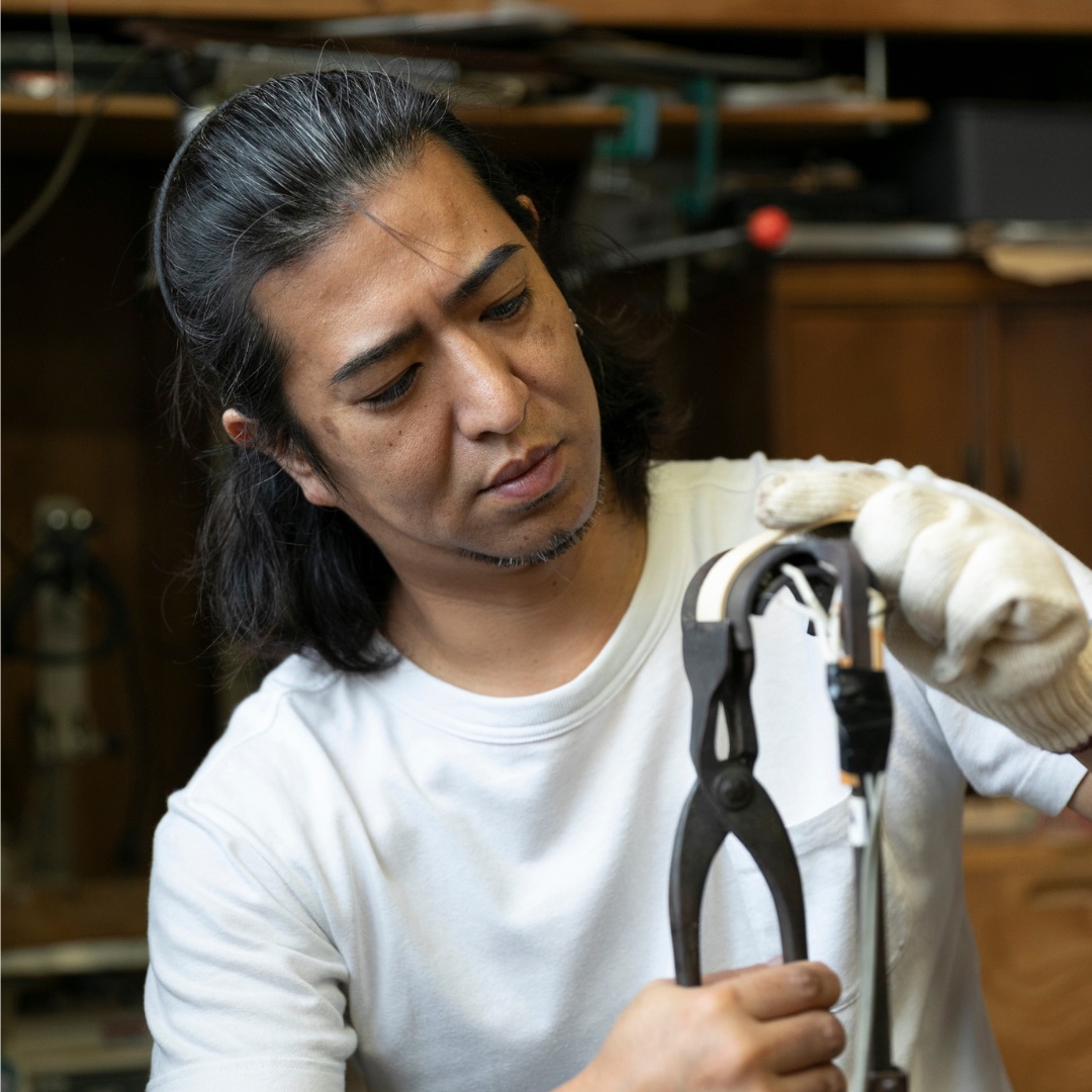 A craftsman in a white T-shirt bends a thin strip with pliers in his workshop