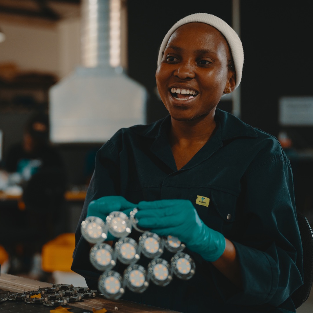 A smiling woman in a work jacket and gloves holds a panel of round LED boards at her workbench