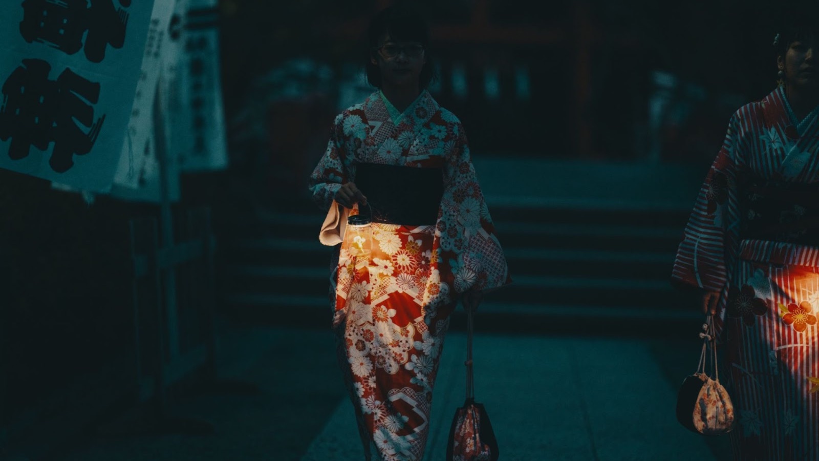 A woman in a patterned kimono walks along a path at dusk, lit warmly from below